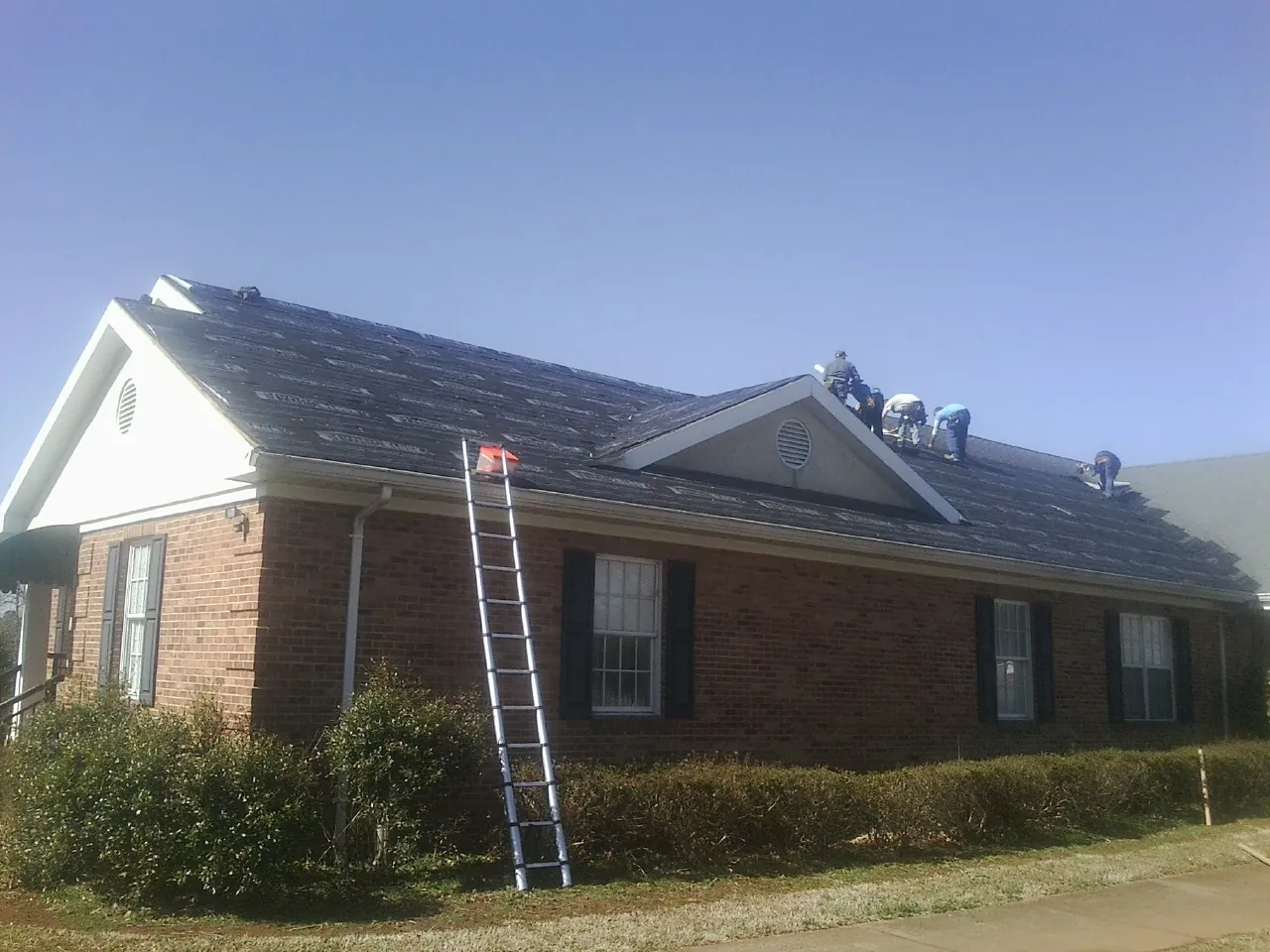 Crew tearing shingles off a brick ranch roof with a ladder set — Emergency Roof Repair work by our crew in Bloxom