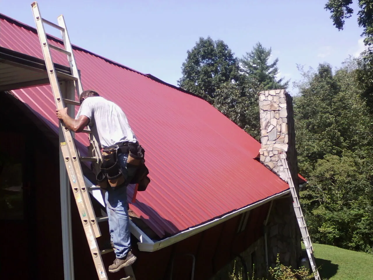 Roofer on a ladder beside a red metal roof and stone chimney — Soffit & Fascia Repair by Roofing Brothers in Bloxom
